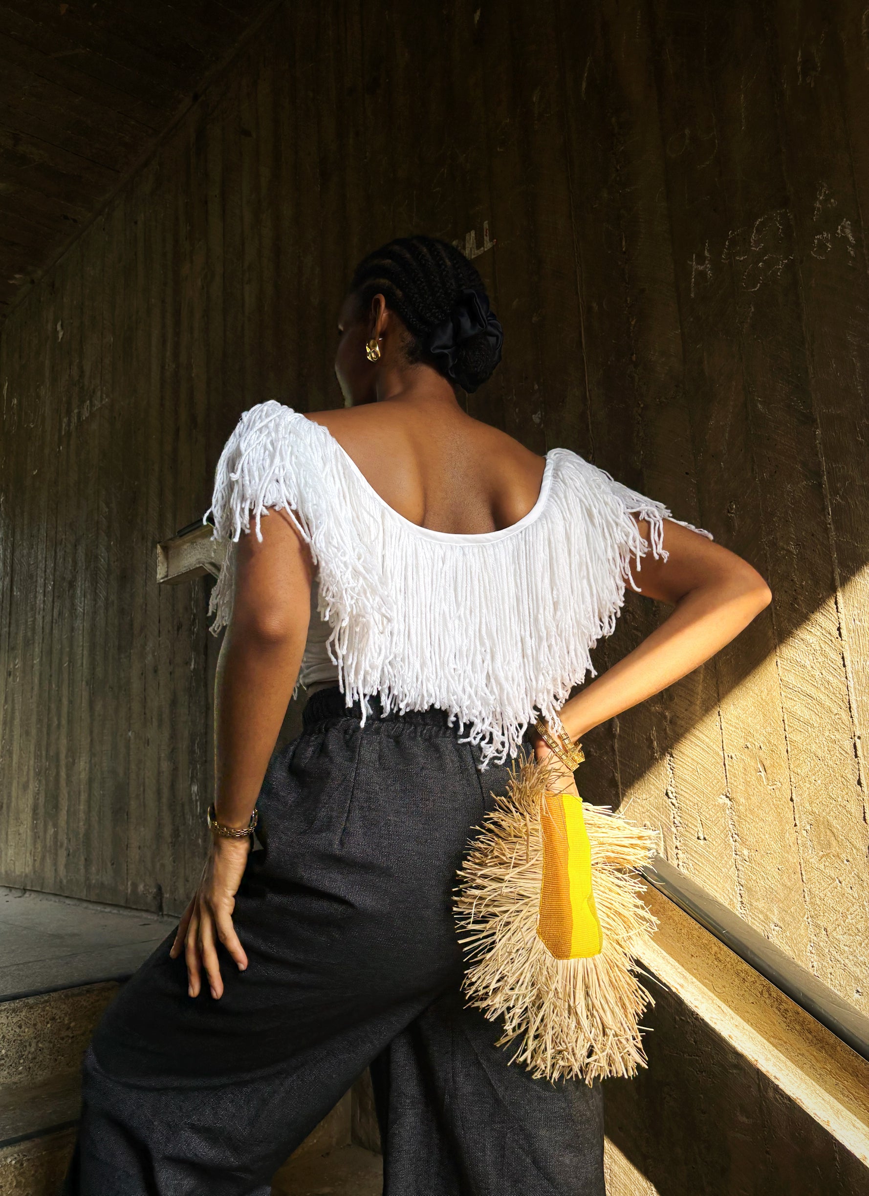 A white fringe top and black pants, holding a yellow and beige bag against a wooden wall.